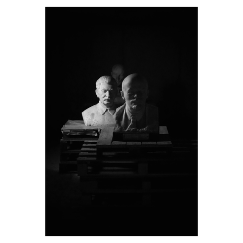This is a portrait format black and white image of three sculptures looming from the darkness of a concrete bunker. Taken at Momento Park in Budapest, the pale stone busts of past Soviet leaders (including Lenin and Stalin) sit on pallets in the darkness in the centre of the frame. A small amount of light from the left highlights the bust of Stalin in the centre-left of the image. The mood is one of decay, gloom and foreboding. Taken with a Sony RX100.