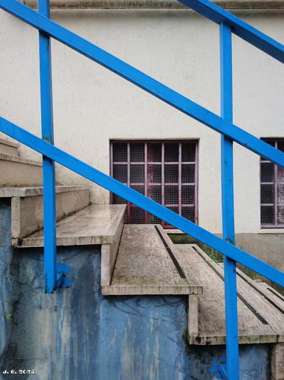 Farbfoto einer von rechts nach links aufsteigenden Treppe, von der Seite aufgenommen. Vor der Treppe ist an der mehr oder weniger blau gestrichenen Mauer ein blau gestrichenes Metallgeländer angebracht. Hinter der Treppe sind in die cremeweiße Mauer zwei vergitterte Fenster eingelassen. Sony Xperia Z5 mit Vignette for Android.