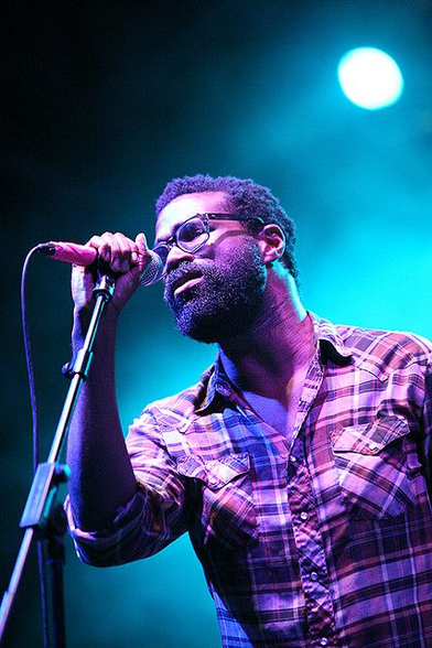 Tunde Adebimpe (born February 25, 1975) is an American musician, singer-songwriter, actor, director, and visual artist, best known as a founding member and co-lead vocalist of the Brooklyn-based band TV on the Radio, with whom he has recorded five studio albums. Here, he is seen leaning into a wired microphone, eyes closed. His beard is gleaming and he’s wearing a comfy flannel shirt, bathed in purple stage lights.