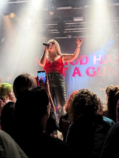 Janine, singer of Vukovi, is performing in front an audience with her band Vukovi. She is wearing a vivid red top and black leather trousers. She holds the mic with her right hand in front of her face, in singing position, while her left hand is raised in a position as if she wanted to show something. Behind her, the background is a black fabric with the name of their latest album "My god has got a gun" written in bold red lettering. In the picture only a couple of the words are visible. In front of her a bunch of heads can be seen. A phone is raised taking a picture of her.