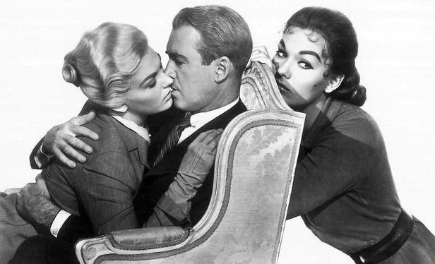 Kim Novak in her characters from Vertigo, for a marketing photo.
In between them is James Stewart, kissing the blonde Kim Novak/Madeleine on the left. He is sitting in a classy rather old seat. On the right, the Judy character (with dark hair) from the movie is behind the seat, looking aside and a bit sad. Also Kim Novak in that role.