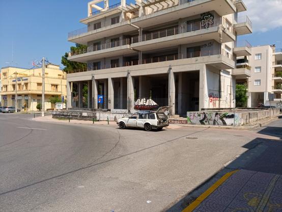 What the toot said. The car is parked in front of an abandoned concrete building construction with Greek graffiti. A yellow building behind flies a Greek flag on top. The asphalt road is grey, blindingly sunny and hot.