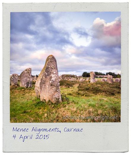 Square photo presented in an old Polaroid frame, showing lines of standing stones in a grassy field on a cloudy evening.