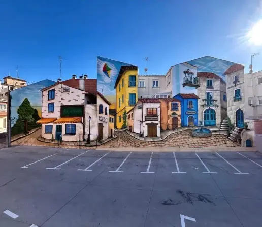 A large trompe-l'œil mural by Rober Bece in Santa Marta de Tormes, Spain, depicting a picturesque town square with cobblestone streets, colorful Mediterranean-style buildings, balconies, a fountain, and a bar with an orange awning. The artwork seamlessly blends with the surrounding architecture, making the entire scene appear three-dimensional. The illusion is especially striking in the way it interacts with the real parking lot in front of it.