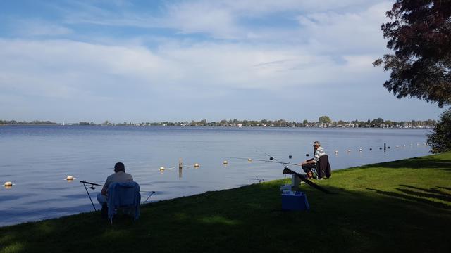 Two casually dressed, older men, sitting five meters apart in camping chairs in the grass at the edge of a lake, while fishing on a sunny day. They are looking out over the lake, their arms on their legs, their fishing poles out over the water resting on holders. On the horizon the opposite shore, an indistinct line of trees and buildings.