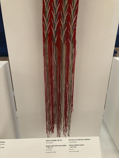 Red, green threads and white seed beads forming a sash that hangs vertically.
Text from display reads:
ONCE-KNOWN ARTIST
Huron-Wendat
Beaded sash with arrow design c. 1800-50
wool and beads
Purchased 1939 (no. 9629)
ARTISTE AUTREFOIS CONNUE
Huronne-Wendat
Ceinture fléchée perlée
v. 1800-1850
laine, perles
Acheté en 1939 (n° 9629)