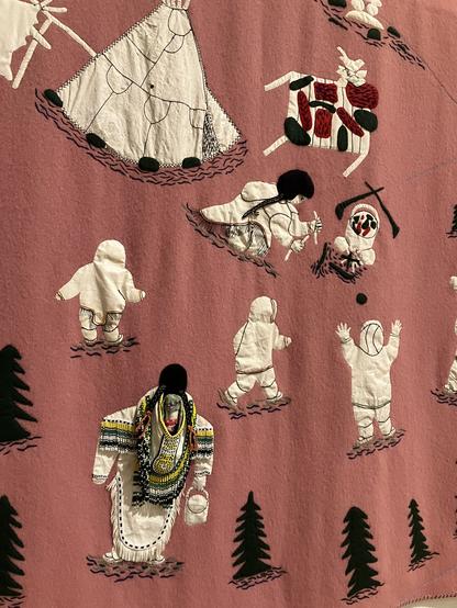 À deep salmon felted wool tapestry featuring Inuit figures in a community with a lodging and central fire. All hand stitched with black threads and coloured beads adding three dimensionality