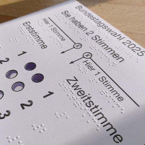 Foto mit schräger Sicht auf ein weißes Kartonpapier mit Schwarz-und Brailleschrift. Schwarze Schrift und Symbole sind ebenfalls erhaben und ertastbar. Oben auf dem Bogen steht: "Bundestagswahl 2025, Sie haben 2 Stimmen". Zwei Pfeile sind jeweils mit "Hier 1 Stimme" beschriftet und weisen nach unten auf zwei Spalten. Die linke Spalte ist mit "Erststimme", die rechte mit "Zweitstimme" beschriftet. In jeder Spalte eine Reihe kreisförmiger Öffnungen, die mit Ziffern nummeriert sind. Im Bildausschnitt stehen die Ziffern 1 bis 3 in Schwarz- und Brailleschrift.