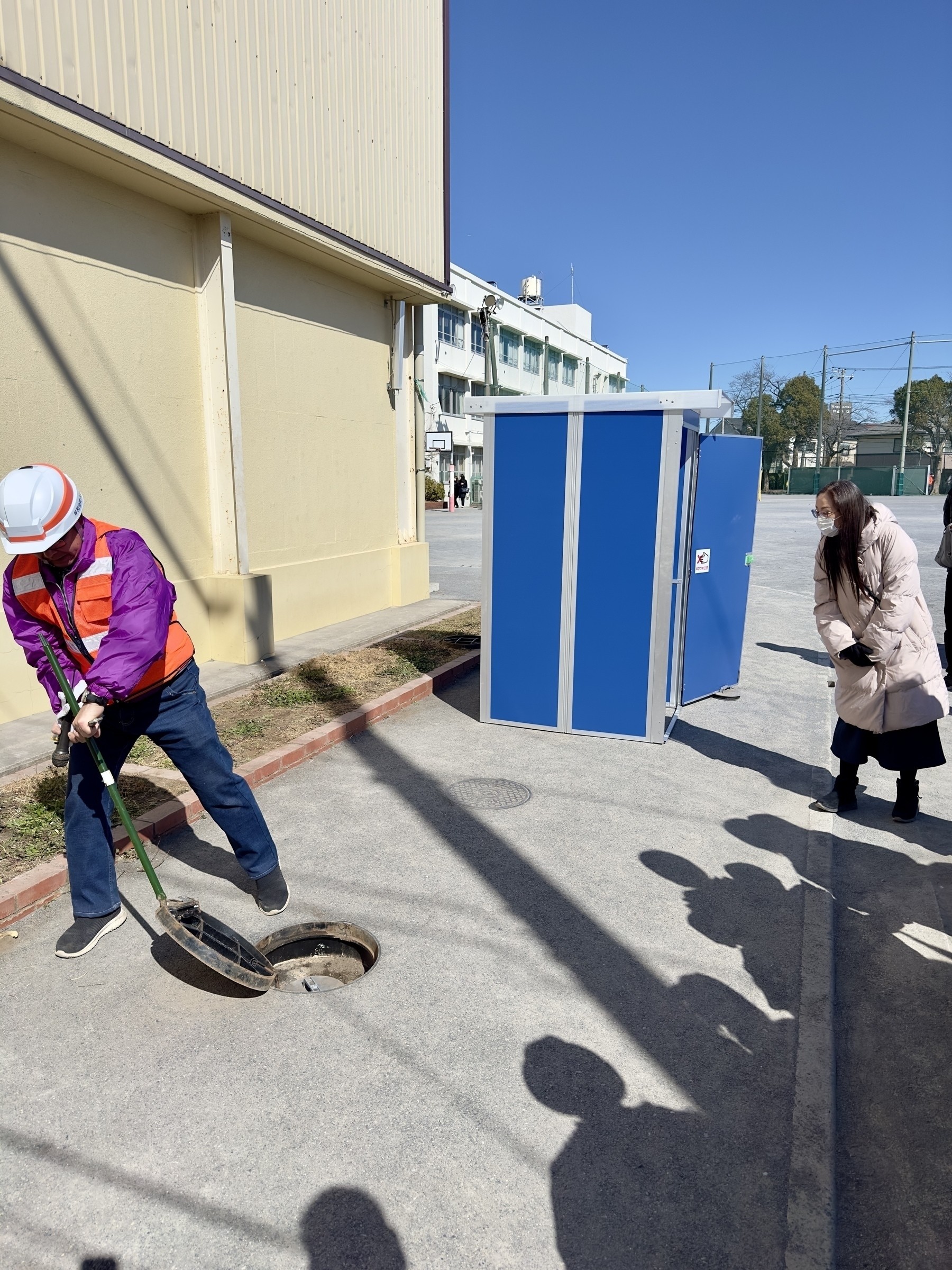 A person in a hard hat and safety vest is using a tool to open a manhole cover on a sidewalk, while another person wearing a mask observes nearby.