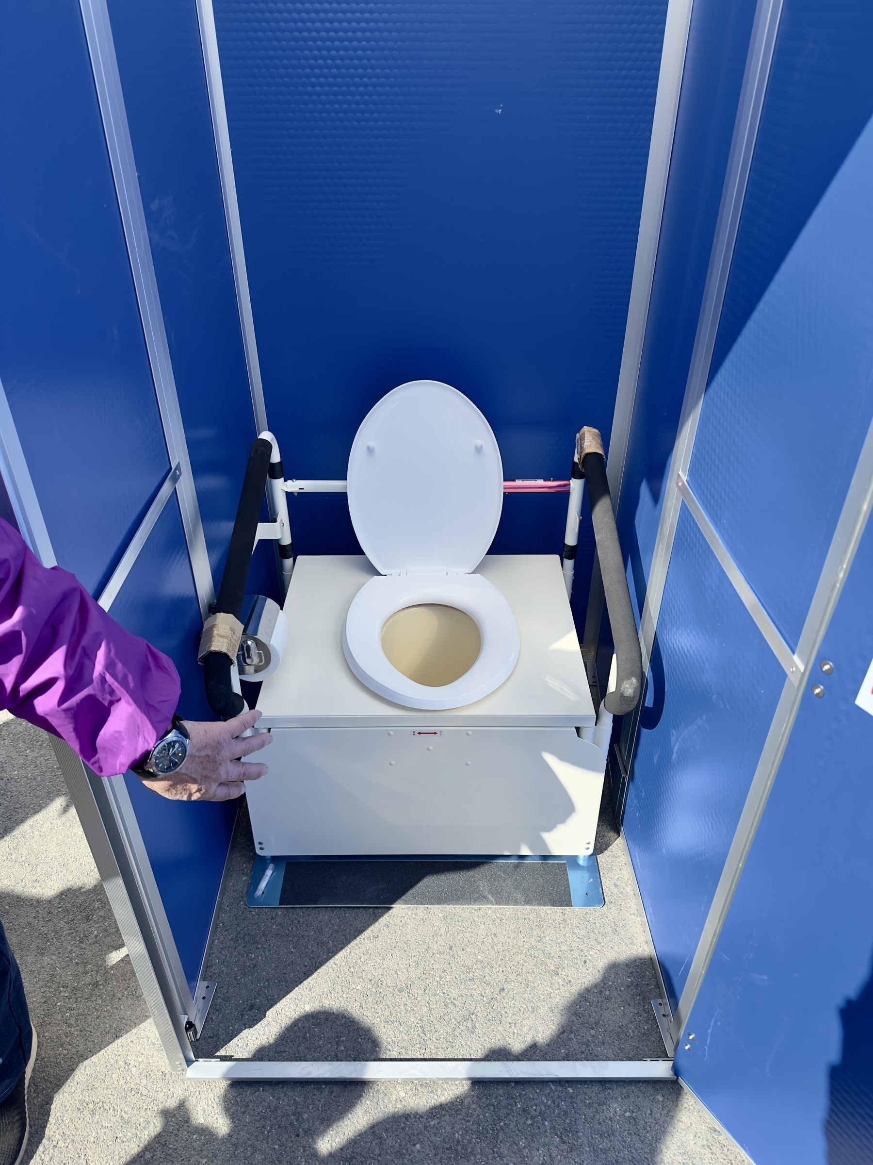 A person is reaching towards an open portable toilet with blue walls and handrails.