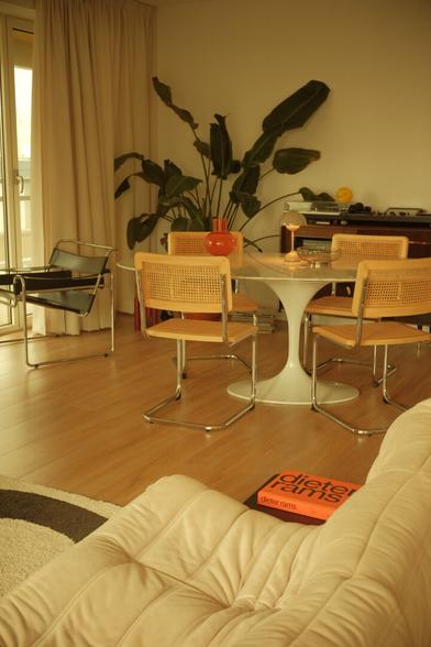 A warmly lit mid-century modern living space featuring a white tulip dining table with four Cesca chairs, a black Wassily chair near a window with long beige curtains, a large green plant, and a vintage wooden sideboard with a turntable and a yellow smiley face ball. In the foreground, a plush Togo chair with a bright orange Dieter Rams book resting on it.
