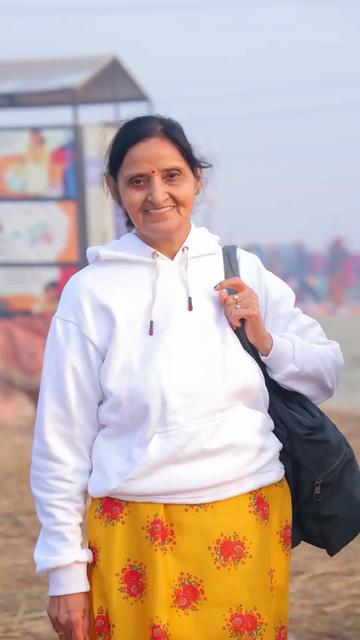 A portrait shot of Sourabh Raaj Jain's mother, smiling as she faces the camera in Prayagraj during the Maha Kumbh Mela. She wears a white hooded sweatshirt over a golden yellow kurta with a red floral pattern, and is carrying a black bag over her shoulder. Her hair is tied back and she is wearing gold rings, one with a large pearl. She wears a red bindi dot painted in the space between her eyebrows where the ajna chakra is found. The image is a still frame from a video that was shared by Sourabh in a post on Instagram on Feb. 3, 2025.