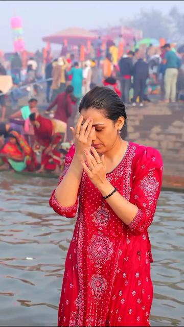 Ridhima ma'am stands in the waters of the Ganges, her eyes closed and hands raised, the third and fourth fingers of her right hand pressed against the space between her eyebrows where the ajna chakra is found. She wears a bright cerise pink long kurta with delicate white embroidery, earrings, a black bead bracelet on her left hand and her wet hair swept backwards smoothly. Behind her, other devotees can be seen on the steps beside the sacred waters in Prayagraj during the Maha Kumbh Mela. The image is a still frame from a video that was shared by Sourabh Raaj Jain in a post on Instagram on Feb. 3, 2025.