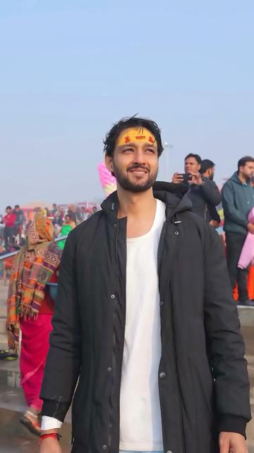 Sourabh Raaj Jain stands facing the camera on the banks of the Ganges, in Prayagraj during the Maha Kumbh Mela. He wears a long white t-shirt and a long padded black jacket, and red and black bands on his right wrist. Tumeric paste has been applied to his forehead, with red lettering and symbols painted on with kumkum. Other devotees fill the steps behind him. He looks towards the low morning sun, which fills the scene with golden light. The image is a still frame from a video that was shared by Sourabh in a post on Instagram on Feb. 3, 2025.
