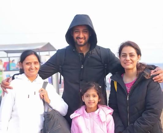Sourabh Raaj Jain stands facing the camera with his arms around his mother, to his left, and Ridhima ma'am, to his right, with Hrishika in front of them. Sourabh wears a long black padded jacket with the hood pulled up and a black rucksack over his shoulders. His mother wears a brilliant white hoodie and is carrying a dark gray-black bag. Ridhima ma'am wears a padded black jacket over a bright cerise kurta with white embroidery. Hrishika wears a light pink padded jacket and pink clips in her hair. They are all smiling. The image is a still frame from a video of their visit to the Ganges, in Prayagraj during the Maha Kumbh Mela, that was shared by Sourabh in a post on Instagram on Feb. 3, 2025.