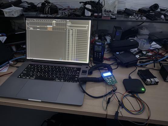 A cluttered workspace featuring a laptop displaying a software interface with a timeline and data logs. There is a handheld cbradio, a mobile cb radio, and a circuit board with illuminated buttons connected via cables. Additional electronic components and tools are visible in the background.