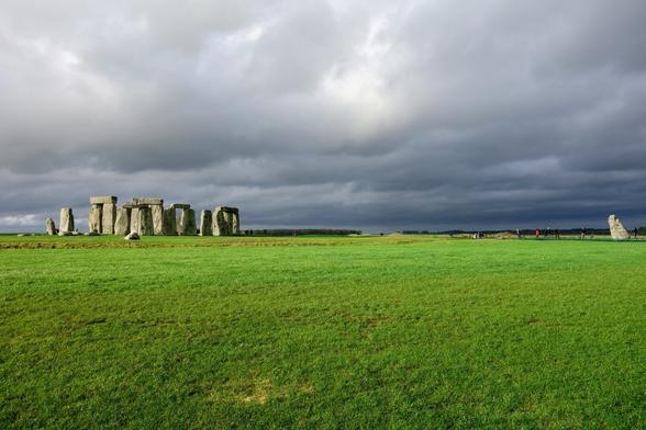A wide shot of Stonehenge under a cloudy sky, with a large green field in the foreground.