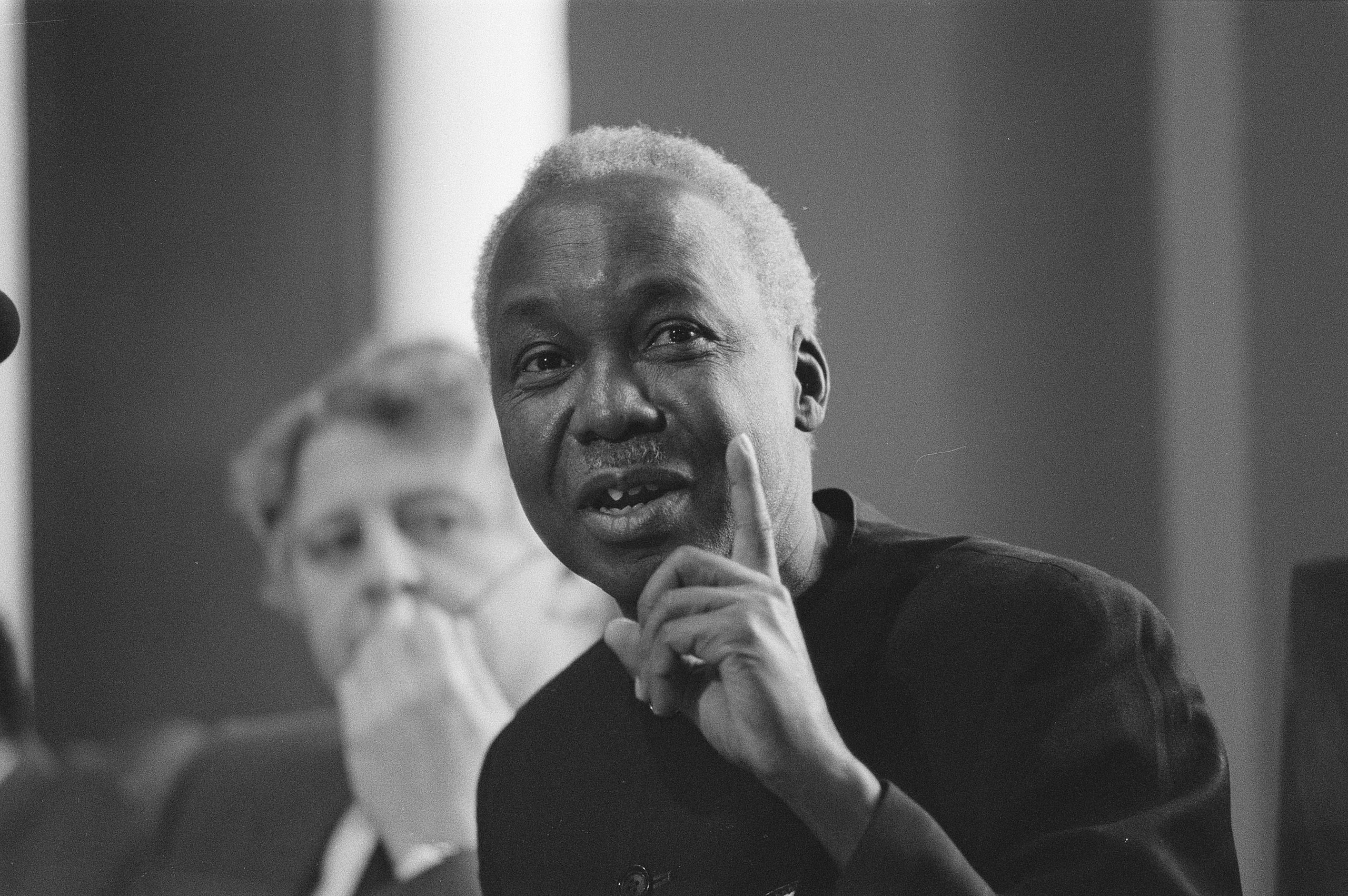 A black man with grey, short hair holds up a finger while in the background, a white man looks on.