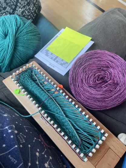 View of a wooden Kb sock loom which has teal and purply-pink yarn on it. A cake of the purply yarn, a colourwork chart, and the teal cashmere yarn are also on the lap.