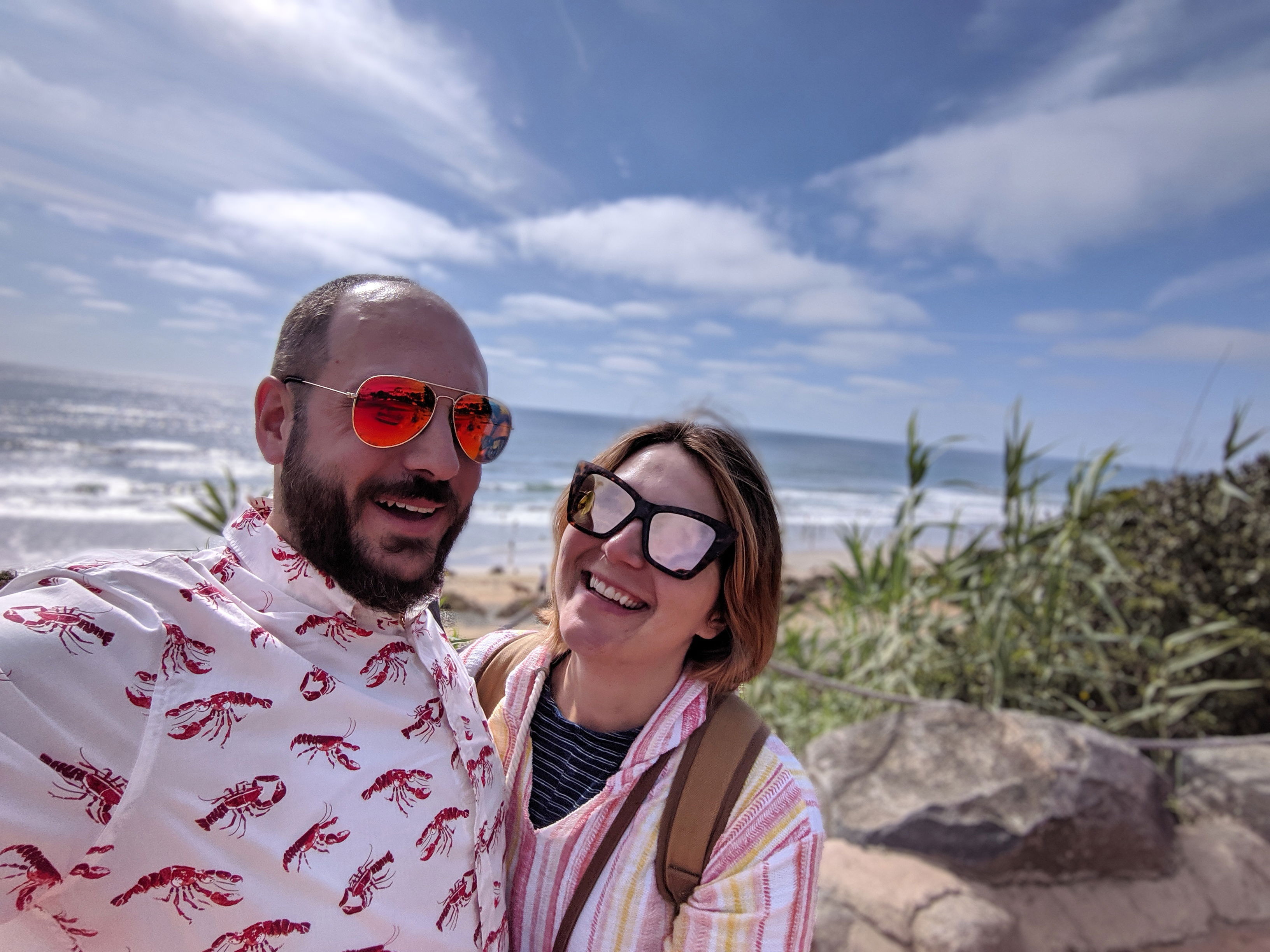 Photograph of @evs and his partner. Both are smiling on the beach. He is wearing a white collared buttondown shirt with a lobster print and mirrored aviator sunglasses. She is wearing a pastel coverall, brown backpack, and mirrored sunglasses.