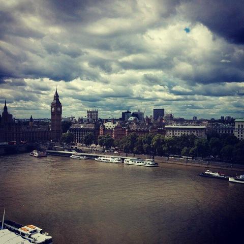 A wide-angle shot of London, taken from an elevated position, featuring the River Thames with several boats and ferries. The Palace of Westminster, including Big Ben, is prominently visible on the left. The city skyline stretches across the background, with various buildings and trees, all under a dramatic, cloudy sky with a small patch of blue.