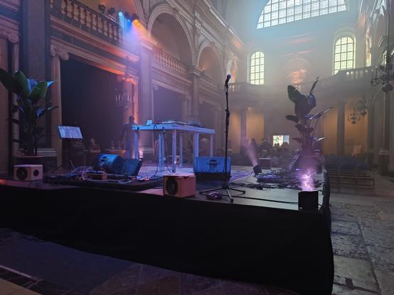Photographie numérique couleur en format paysage.
Dans une église, une scène au centre, avec sur une table 2 platines, une table de mixage et des piles de vinyles. Au sol, des amplis et des plateaux de claviers  pédales d'effets sonores pour les concerts suivants.
L'ambiance est enfumée et douce. 2 plantes vertes sont posées à 2 angles de la scène et éclairées par dessous.