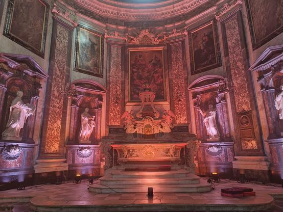 Photographie numérique couleur en format paysage.
Le chœur d'une église baroque richement décoré et éclairé dans des tons orange et rose.
Des marchés arrondies précèdent l'autel.
