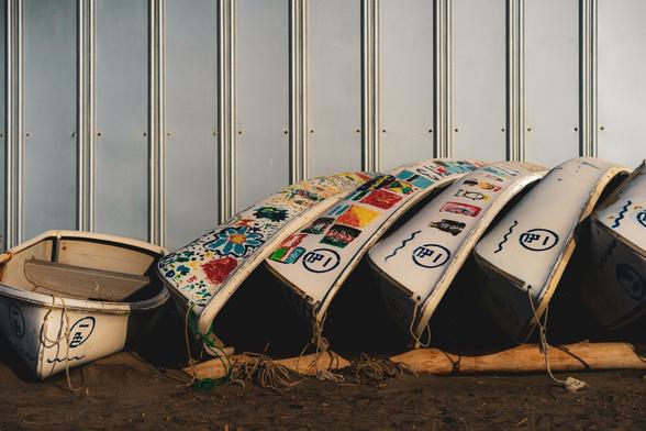The image shows several rowboats propped against a wall. Some of them have colorful, painted designs on their undersides. There are ropes tied around the boats, and the scene appears to be outside, possibly at a beach or near a waterfront.