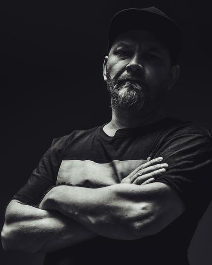The image is a moody, black and white portrait of a person with a beard, wearing a hat and a T-shirt. The person has their arms crossed and is looking directly at the camera. The lighting is dramatic, with shadows emphasizing their features.
