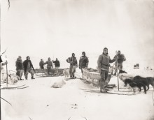 A historical photo of a number of men in heavy winter clothing pose on skis and with dog sleds on a vast stretch of icy landscape. Public Domain.