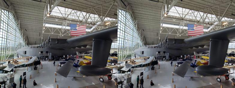 Stereoscopic, parallel view. Interior of an air museum, looking at a the underside of the left wing of a very large seaplane which absolutely dwarfs the other exhibits. It has a boat-like body, pontoons coming from below the wing tips, and is painted military grey.