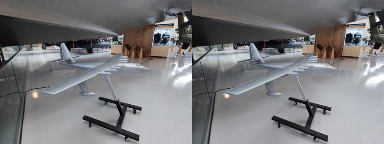 Stereoscopic, parallel view. Interior of an air museum, looking at a model of a seaplane. It has a boat-like body, pontoons coming from below the wing tips, is painted a silvery pewter. Perhaps a 1.5 meter wingspan? It is lacking in details to show the overall shape of the plane. It is close in under the body of the actual plane above it.