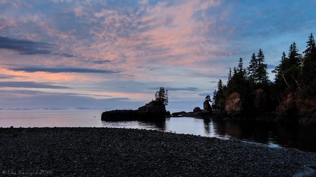 A very dark shore, small stones, curves from the bottom as it meets the calm water. The tide is out and there is a small rock island with a single small group of evergreens on the right side. There are other rock formations out into the water from a dark shore with trees at the top of the low cliff. The sunset is a muted pink and darker blue in a very blue sky and in the distance across the bay, it's either misty or low hills, which is hard to make out.