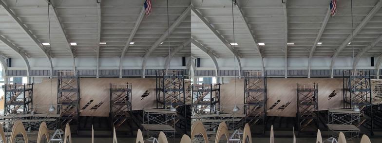 Stereoscopic, parallel view. A diorama of the construction of the Spruce Goose seaplane. We are inside a recreation of the hanger in which it was built. This side view shows the side of the Goose, with the nose on the left and the body toward the right. The nose of the plane has been partially primed white and then painted grey, but where it isn't painted, we can see how the planks were laid along the surface.The leading edge of ribs of the wing under construction run along the bottom of the image.