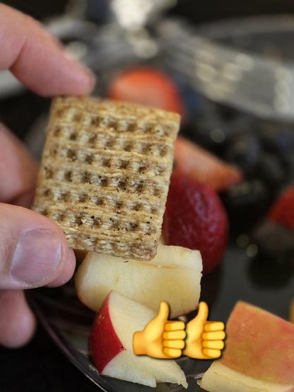 Photo of a hand holding a single triscuit square cracker, with a plate of fruit out of focus in the background.