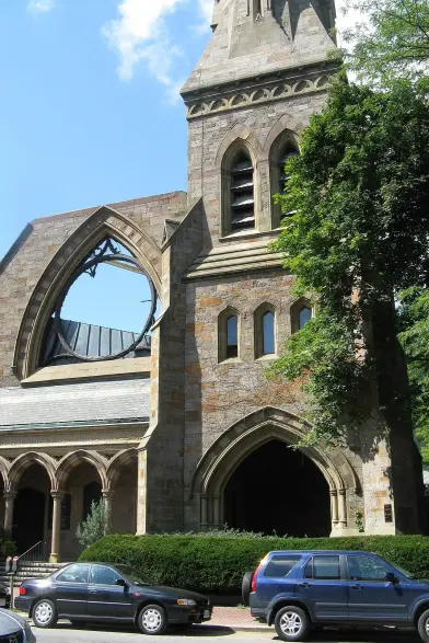 The image depicts a stone building that appears to be a church or a similar structure, characterized by Gothic architectural elements, such as pointed arches and a bell tower. The building shows signs of damage, notably a large hole in the side, possibly indicating that it is in a state of disrepair. Surrounding the building, there are trees and parked cars in the foreground, under a clear blue sky.

Image Credits: Wikimedia / John Phelan / CC BY-SA 3.0