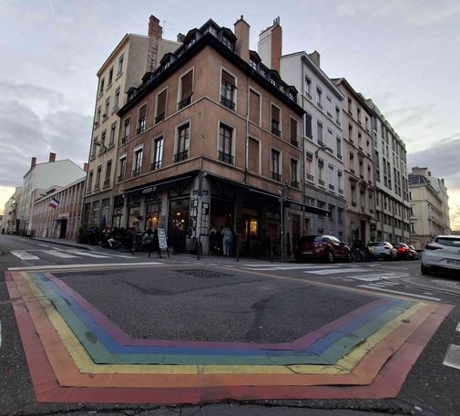 un carrefour de rues dans le 7e arrondissement de Lyon, quartier de la Guillotière où un arc-en-ciel relie les passages pietons. en angle un immeuble ocre avec au pieds un bar assez connu du quartier le Livestation DIY
