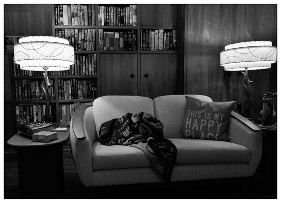 The image depicts a cozy reading nook in a black and white format. It features a small, cozy sofa adorned with a cushion that reads "THIS IS MY HAPPY PLACE" and a patterned throw blanket. Next to the sofa, there is a small table with a few books stacked on it, suggesting it is used for reading. Behind the sofa is a built-in bookshelf filled with numerous books, creating a backdrop that emphasizes a love for reading. The area is illuminated by two stylish table lamps with distinctive round, ringed shades, adding a soft and inviting ambiance to the space. The overall setting suggests a tranquil and intimate space perfect for relaxation and reading.