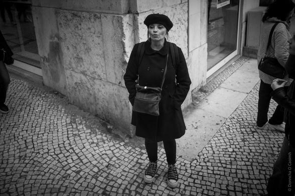 A black and white street photograph of a woman in a beret and coat standing on a cobblestone street.