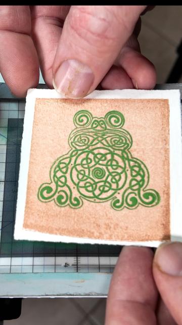 Fingers hold up a small (7cm square) print of a knotwork frog in green on a copper background on toothy white paper.