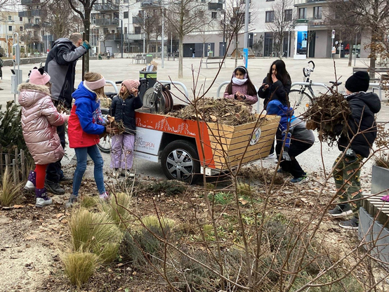 Im Vordergrund liegen Beete die sich noch im Winterschlaf befinden. Viele jüngere Kinder sammeln altes Laub aus den Beeten ein und bringen es zu einem Lastenrad mit großem Aufbau