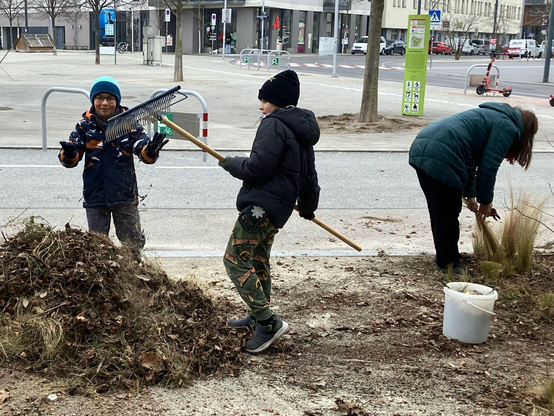 Zwei Kinder mit Mützen und Winterjacken stehen bei einem Laubhaufen in der Seestadt Lounge. Ein Kind hat einen Rechen in der Hand. Im Hintergrund schneidet eine Frau in einem grünen Mantel eine Pflanze. Im Hintergrund sieht man den Hannah-Arendt-Platz.