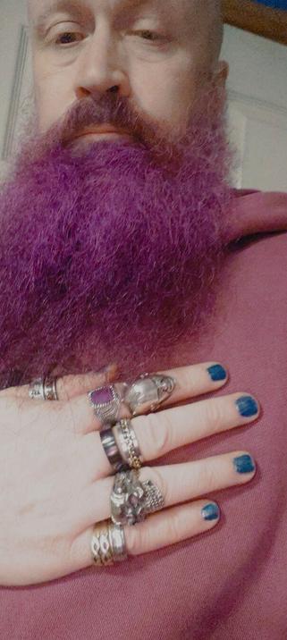 A photo showing my face and upper chest. My hand, wearing multiple rings on several fingers, it's resting on my chest. I'm wearing blue nail polish on my fingernails. Some of the rings are statement pieces that cover a significant portion of the finger, while others are simpler bands. My beard is purple.