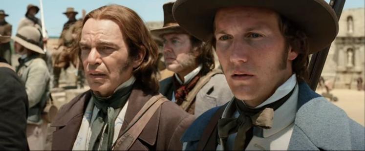 Two men in early American Victorian-era clothes and hair, with coats and high collars. They're standing amidst other men in various dress, some more rough and cowboy-style than others. Both men are looking out toward something offscreen and seem worried.