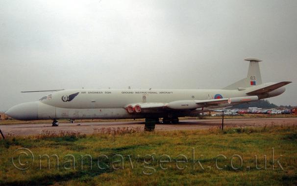 Side view of a grey, 4 engined military jet patrol aircraft parked facing to the left.
The plane's upper fuselage is a dark grey, the lower fuselage and the bulbous nose and tail-coner are a lighter grey.
The engines are buried in pairs in the wing root, with red covers over the inlets.
There are black "Air Engineer Sqn" and "Ground Instructional Airframe" titles on the upper fuselage in black.
There is a red and blue roundel on the rear fuselage, and a red and blue fin flash on the tail, under the number "63".
Green grass fills the foreground, with cars parked in a field in the background, visible under the planes tail, under grey skies.