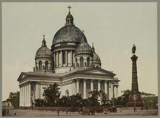 The image shows a large, ornate cathedral with multiple domes and columns. The architecture is grandiose and intricate, featuring classical elements such as Corinthian columns, pediments, and decorative cornices. Atop the main dome sits an Orthodox cross, signifying its religious purpose.

In front of the cathedral stands a tall monument or column, which appears to be dedicated to military honors given the presence of figures resembling soldiers on top. The surroundings depict a street scene with horse-drawn carriages parked along the side and pedestrians walking by.

The overall atmosphere conveys a sense of historical significance and architectural beauty typical of early 20th-century Russian cityscapes, specifically in Saint Petersburg during that period.