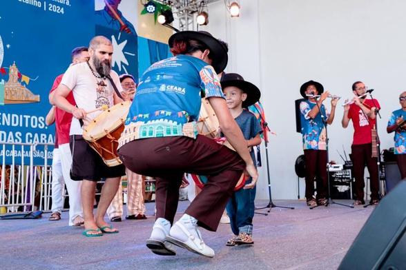 Pessoas em um palco dançando e tocando pife e zabumba. Uma dessas pessoas é uma criança.