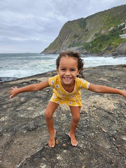 A young girl with curly brown hair stands on a rocky outcrop by the sea, smiling widely with her arms outstretched. She is wearing a short-sleeved yellow dress with a small white floral pattern. The sea behind her is choppy with whitecaps, and a large, green, rocky hill dominates the background under an overcast sky.