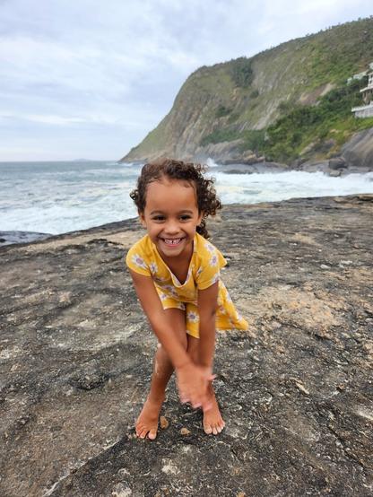 A young girl with curly brown hair stands on a rocky shoreline, beaming at the camera. She is wearing a yellow dress with a white floral pattern and is barefoot. She is playfully leaning forward with her hands clasped in front of her. Behind her, waves crash against the rocky shore, and a large, green hill rises in the distance under a cloudy sky.