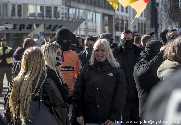 nazidemo, im vordergrund drei blonde frauen, zum teils vermummt, dahinter eine person in orangener jacke und vollvermummt mit zweilochsturmhaube